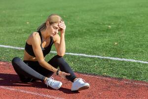moe vrouw atleet in sportkleding zittend Aan een rennen spoor, Holding haar hoofd in spanning of uitputting na opleiding. foto