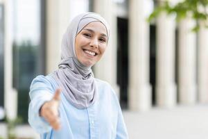 een vrolijk moslim vrouw vervelend een hijab strekt zich uit een handdruk, symboliseert Welkom en overeenkomst, met een wazig gebouw achtergrond. foto