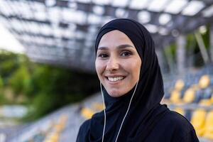 portret van gelukkig moslim vrouw in hijab en hoofdtelefoons Aan rechtbank, aan het doen sport- en luisteren naar muziek, glimlachen en op zoek Bij camera. foto