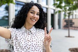 vrolijk jong vrouw met gekruld haar- nemen een selfie, knippert een vrede teken buitenshuis, uitdrukken vreugde en positiviteit. foto