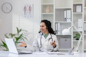 een professioneel vrouw dokter met een stethoscoop verloofd in een interactief online overleg, glimlachen hartelijk in een goed uitgerust medisch kantoor. foto