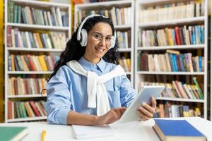 een vrolijk vrouw vervelend hoofdtelefoons is comfortabel gebruik makend van een digitaal tablet in een goed gevuld bibliotheek, omringd door talrijk boeken. foto