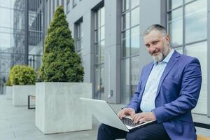 senior knap geslaagd mannetje zakenman in pak werken met laptop online. hij zit Aan een bank in de buurt de kantoor centrum, looks Bij de camera, glimlacht. foto