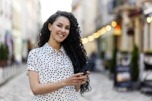 portret van een jong mooi Latijns Amerikaans vrouw wandelen in de avond stad, Holding een telefoon, glimlachen en op zoek Bij de camera. foto