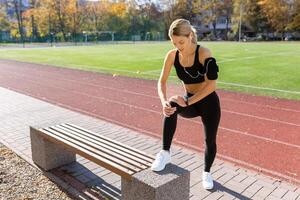 fit vrouw uitrekken been Aan bank Bij zonnig bijhouden veld, uitstralend Gezondheid en vitaliteit, vervelend sportkleding, oortelefoons foto