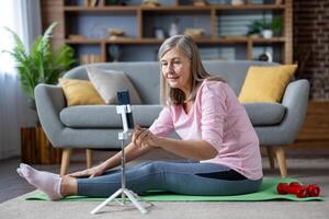 senior volwassen grijs haren vrouw aan het doen geschiktheid zittend Aan oefening mat Bij huis in leven kamer Aan vloer, gebruik makend van statief statief met telefoon, aan het kijken online klasse en opleiding . foto