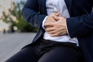 handen van een jong Mens in een bedrijf pak Holding zijn maag, zittend buiten Aan een bank. detailopname foto. foto