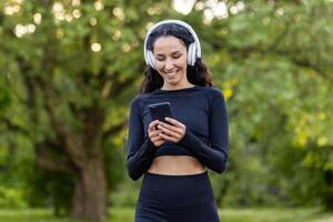 geslaagd slank Latijns Amerikaans vrouw in park gedurende actief oefening en jogging toepassingen smartphone app, sportvrouw glimlachen in hoofdtelefoons luisteren naar audio podcasts en online radio. foto