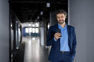 volwassen ervaren geslaagd zakenman in bedrijf pak wandelen binnen kantoor gebouw, Mens wandelen naar beneden gang met telefoon in handen, baas gebruik makend van app smartphone, lezing en glimlachen tevreden foto