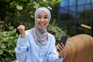 jong mooi moslim vrouw in hijab zittend Aan bank buiten kantoor gebouw, zakenvrouw op zoek Bij camera, ontvangen online bericht, zege succes, Holding hand- omhoog vieren triomf foto