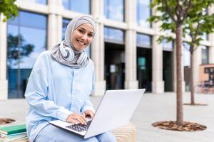 een vrolijk moslim vrouw vervelend een hijab zit buitenshuis met een laptop, weergeven produktiviteit en connectiviteit in een stedelijk instelling. zij is werken in de buurt modern kantoor gebouwen en bomen. foto