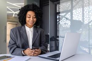 geslaagd zakenvrouw Bij werkplaats binnen kantoor gebruik makend van telefoon, vrouw arbeider lezing bericht en lachend, Afrikaanse Amerikaans vrouw Holding smartphone gebruik makend van online sollicitatie. foto