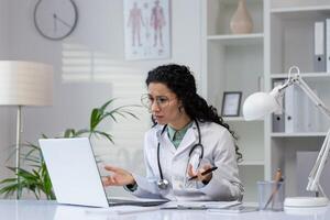 een professioneel latino vrouw dokter is gefocust Aan haar werk, overleg plegen medisch informatie Aan haar laptop in een modern kliniek. foto