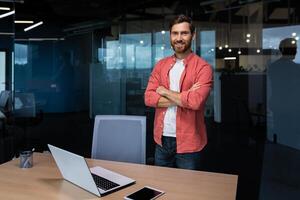 portret van een geslaagd zakenman binnen de kantoor, een Mens met gekruiste armen glimlacht en looks Bij de camera, een werknemer staat in de buurt een werkplaats met een computer in een rood overhemd en bril. foto