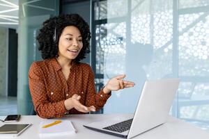 jong mooi vrouw pratend naar klanten van een afstand, Afrikaanse Amerikaans vrouw arbeider glimlachen gebruik makend van koptelefoon telefoon en laptop voor online overleg, klant onderhoud. foto