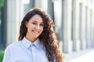 portret van een zelfverzekerd en geslaagd jong Latijns Amerikaans vrouw met gekruld haar- buiten een stad kantoor gebouw. foto