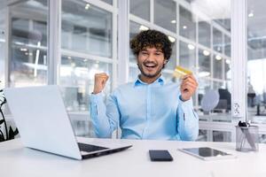 jong spaans Mens zittend in de kantoor Bij de bureau, Holding een credit kaart op zoek Bij de camera en gelukkig. shows een zege gebaar van succes met zijn handen. foto