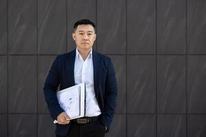 portret van een jong geslaagd echt jong Aziatisch Mens staand buiten een bedrijf centrum tegen een muur, Holding documenten en een overeenkomst in zijn hand. hij looks ernstig in de camera. foto