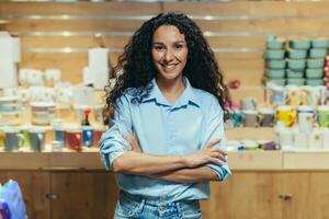 portret van bedrijf baasje, spaans vrouw beheren geschenk winkel, vrouw met gekruld haar- op zoek Bij camera en glimlachen met armen gekruiste foto