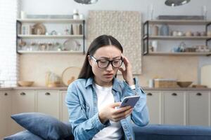 bezorgd jong Aziatisch vrouw houdt de telefoon in haar handen, ontvangen slecht nieuws door bericht. zittend Bij huis Aan de bank, Holding haar hoofd, zenuwachtig. foto