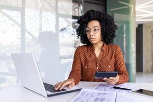 echt denken vrouw arbeider financier werken met documenten en papieren binnen kantoor Bij werkplek, zakenvrouw controle rapporten, gebruik makend van rekenmachine en laptop in papierwerk. foto