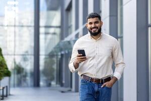 portret van een glimlachen jong nidhi Mens wie is lachend, Holding zijn hand- in zijn jeans zak, gebruik makend van een mobiel telefoon en op zoek vol vertrouwen in de camera foto