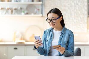 online winkelen. een glimlachen jong mooi Aziatisch vrouw maakt een online boodschappen doen bestellen en pays met een zonder contant geld account. hij is Holding een telefoon en een credit kaart. zittend Bij huis in de keuken. foto