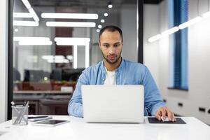 een professioneel individu diep gefocust Aan een taak, werken efficiënt Aan een laptop in een goed verlicht modern kantoor instelling. foto