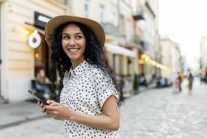 een mooi jong vrouw wandelingen avond stad in een hoed, een glimlachen Latijns Amerikaans vrouw houdt een smartphone in haar handen. een toerist met gekruld haar- types een bericht en bladert online Pagina's Aan de telefoon. foto