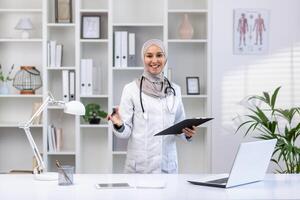 aantrekkelijk Islamitisch medisch professioneel poseren met map tablet in kabinet met portable computer. gelukkig vrouw dokter gevoel tevreden met controle resultaten en maken apotheek recept. foto