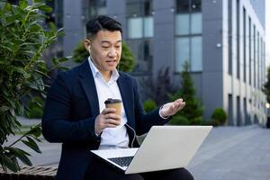 een goed gekleed zakenman bezighoudt in een telefoontje gebruik makend van zijn laptop buiten een kantoor gebouw terwijl Holding een koffie beker. foto