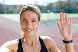 vrouw atleet na jogging in stadion pratend met vrienden en opname online blog, volwassen blond met hoofdtelefoons op zoek Bij smartphone camera lachend, telefoontje met koptelefoon. foto