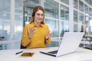 volwassen volwassen bedrijf vrouw met blond haar- werken binnen licht kantoor, klant onderhoud tech arbeider glimlachen en pratend naar klant overleg plegen, gebruik makend van koptelefoon en laptop voor afgelegen telefoongesprek. foto