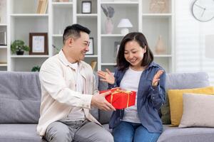 gelukkig Aziatisch familie Mens en vrouw samen Bij huis zittend Aan sofa in leven kamer, man geven geschenk naar vrouw in viering, paar knuffelen en gelukkig. foto