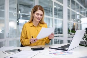 een aantrekkelijk blond vrouw werken in een modern kantoor, zit Bij een bureau in voorkant van een notitieboekje, doet papierwerk, looks aandachtig Bij de computer scherm, jota naar beneden informatie. foto