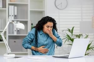 een volwassen vrouw ervaren misselijkheid Bij haar bureau met een hand- over- haar mond en Holding haar maag, mogelijk wijzend op ziekte of ongemak. foto