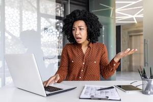 ontevreden en teleurgesteld verdrietig vrouw Bij werkplaats op zoek Bij laptop, lezing online e-mail kennisgeving, zakenvrouw van streek en verward zittend binnen kantoor. foto