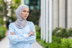 een professioneel moslim vrouw staat vol vertrouwen in een blauw kleding met een hijaab, armen gekruist, tegen een stedelijk backdrop met groen en modern architectuur. foto