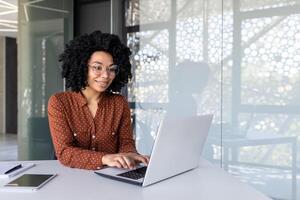 gelukkig en glimlachen Afrikaanse Amerikaans zakenvrouw typen Aan laptop, kantoor arbeider met gekruld haar- en bril gelukkig met prestatie resultaten, Bij werk binnen kantoor gebouw foto