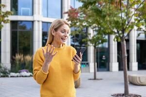 een blij middelbare leeftijd vrouw in een helder geel trui toepassingen haar smartphone, zichtbaar gelukkig over de nieuws ze is ontvangen, staand in een modern stedelijk park. foto