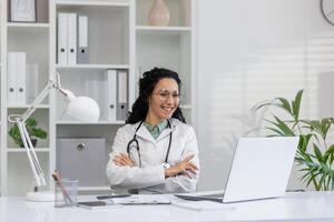 portret van een vrolijk spaans vrouw dokter vervelend bril, zittend in haar kliniek kantoor met een laptop, uitstralend professionaliteit en zorg. foto