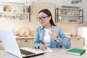 afgelegen werk gedurende quarantaine. jong Aziatisch bedrijf vrouw freelancer werken van huis. zittend Bij een tafel met een laptop, dirigeren een online ontmoeting. conferentie. zegt Hallo naar de camera, schrijven. foto