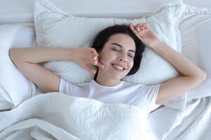 top visie, jong mooi brunette vrouw slapen in bed met ogen gesloten, glimlachen in slaap vervelend wit t-shirt pyjama foto