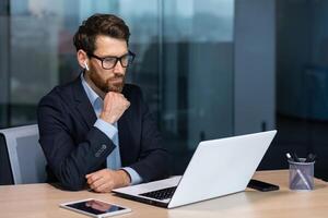 echt denken volwassen zakenman werken binnen modern kantoor gebouw, senior baas in bedrijf pak en bril werken zittend Bij laptop, Mens met baard denken over toekomst strategie plan. foto