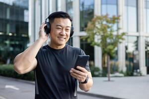 Aziatisch jong Mens in sport- uniform staand in de midden- van de straat vervelend hoofdtelefoons en Holding een telefoon, luisteren naar muziek, audioboek, podcast, aan het doen sport. foto