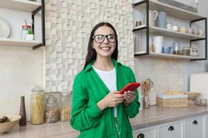 jong mooi vrouw in bril en groen overhemd in de keuken gebruik makend van een rood telefoon foto