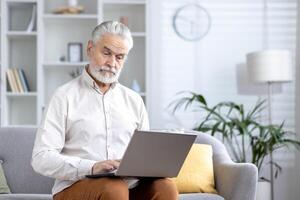 gefocust ouderen heer browsen de internet Aan zijn laptop terwijl zittend Aan een bankstel in een goed verlicht leven kamer. foto