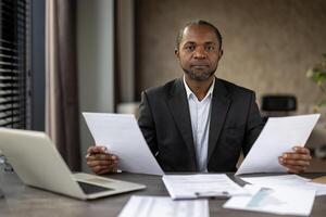 een Mens in een pak is zittend Bij een bureau met papieren in voorkant van hem. hij is Holding twee papieren in zijn handen en hij is gefocust Aan hen. de tafereel suggereert een professioneel instelling foto