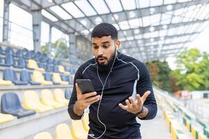 een verbaasd jong Mens in sportkleding looks Bij zijn telefoon met een verontrust uitdrukking Bij een leeg stadion zitplaatsen Oppervlakte. foto