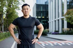 portret van een jong echt Mens, Aziatisch sportman, loper en atleet staand in sportkleding Aan een stad straat, Holding zijn handen door zijn zijden en op zoek Bij de camera. foto